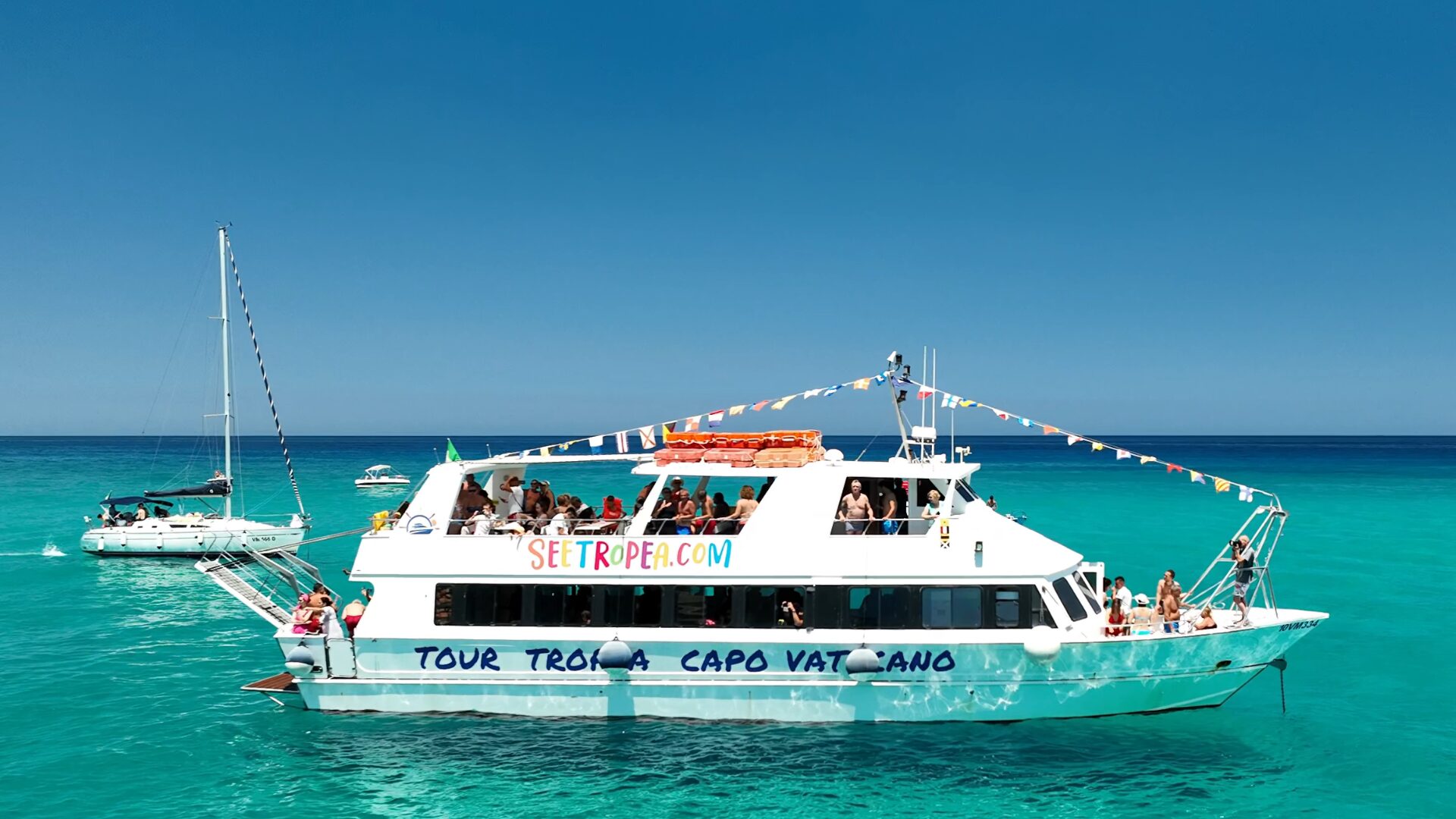 Battello bianco con scritte SEETROPEA.COM e TOUR TROPEA CAPO VATICANO, passeggeri, bandierine, mare turchese